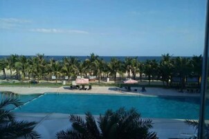 A pool with a view of the ocean, palm trees, and beach umbrellas.