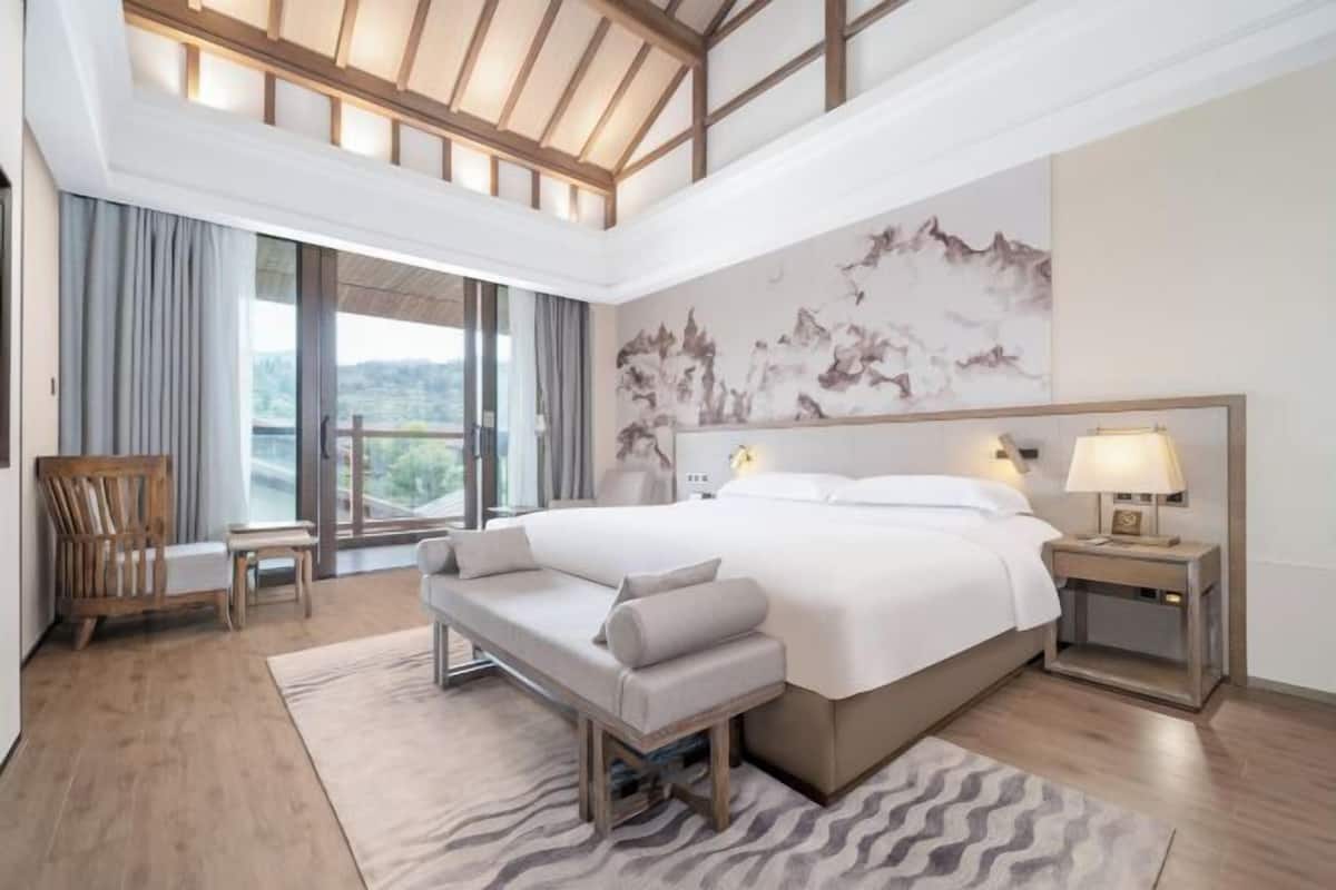 Habitación de hotel moderna con una cama grande, un banco, un escritorio y vistas al exterior.