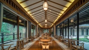 A spacious corridor with wooden tables and chairs, large windows, and a wooden ceiling with hanging lights.