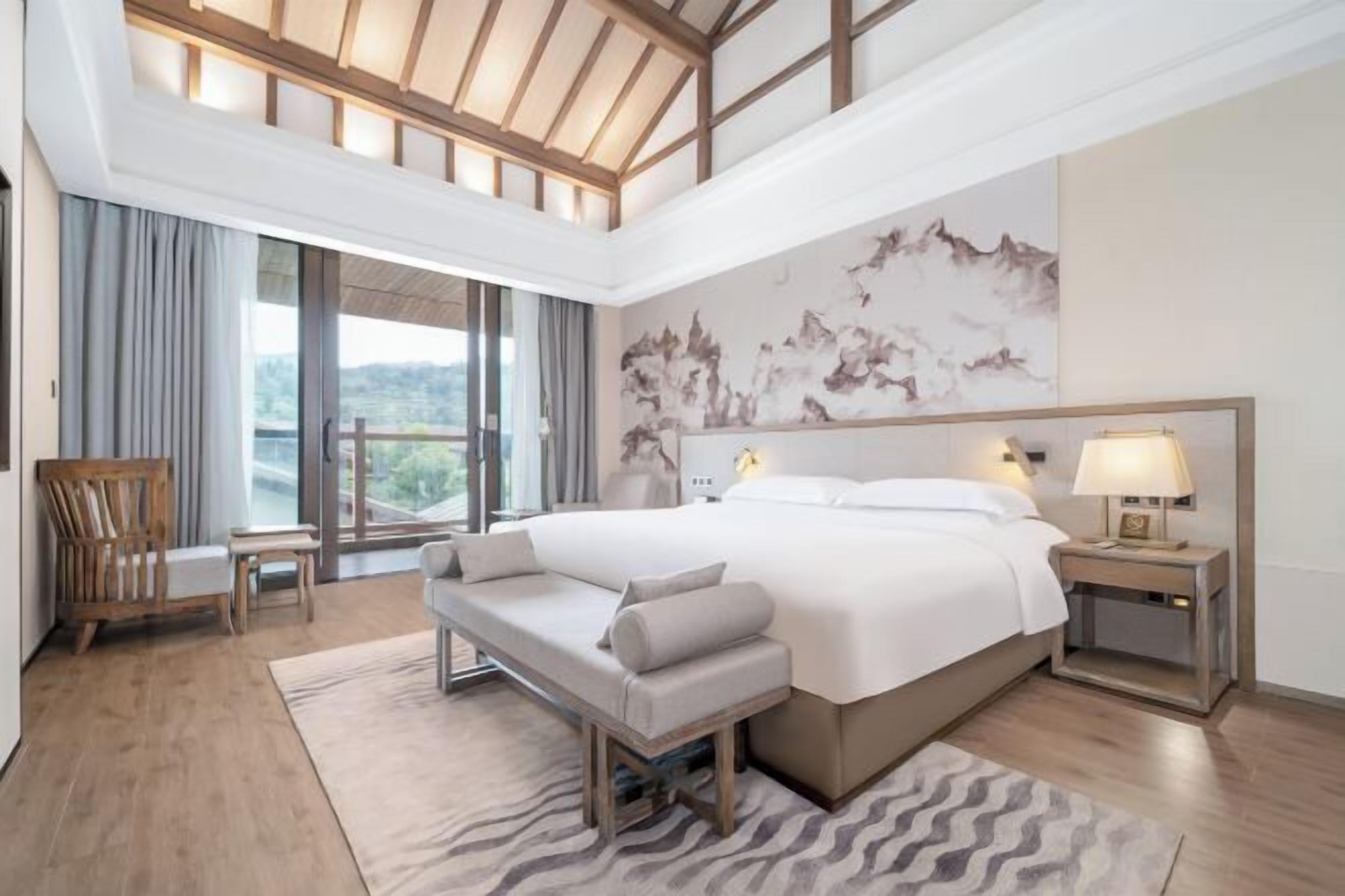 A modern hotel room with a large bed, a bench, a desk, and a view of the outdoors.