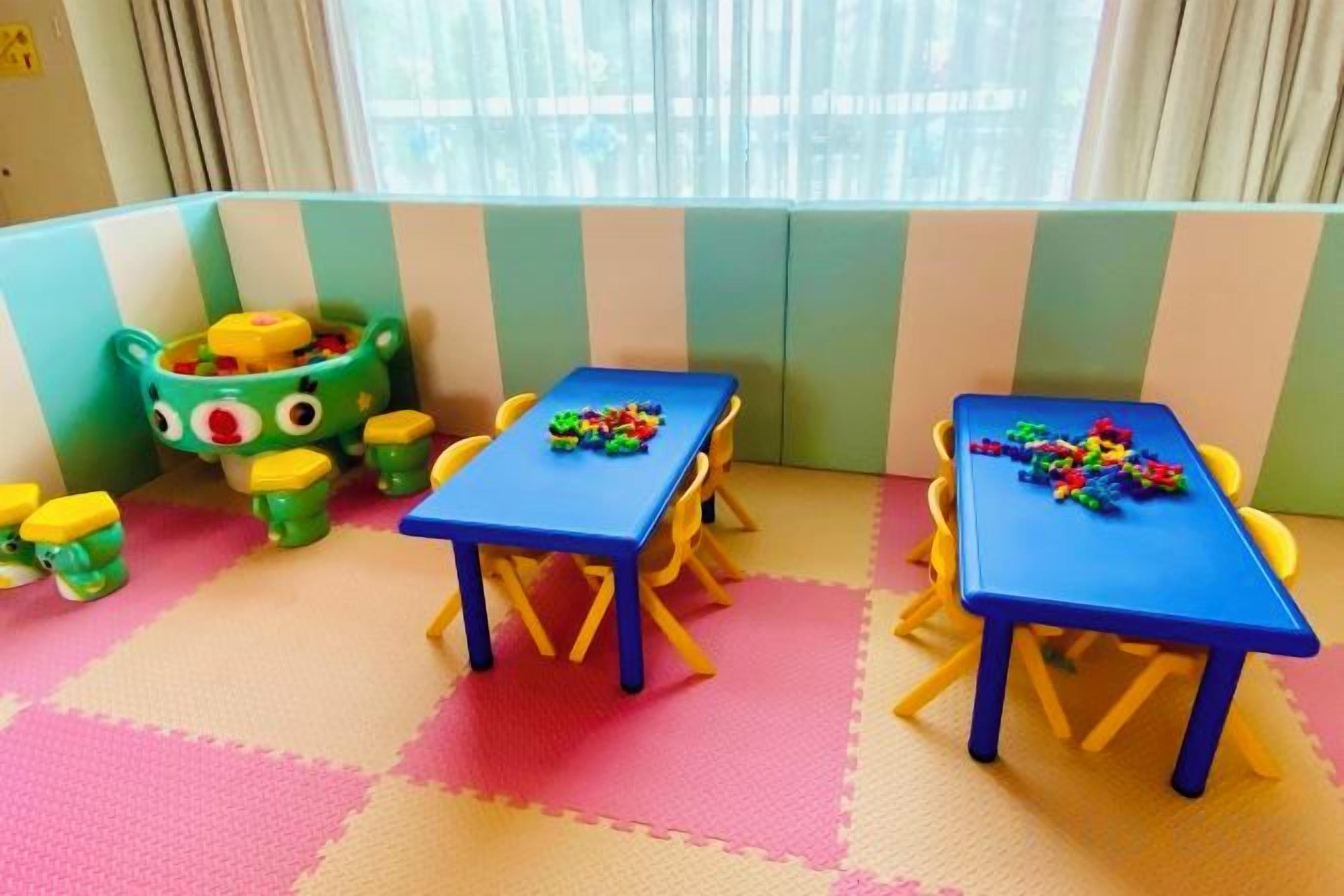 Ein Spielbereich für Kinder mit zwei Tischen und Stühlen, einem grünen Spielzeugschrank und einem Fenster mit Vorhängen.