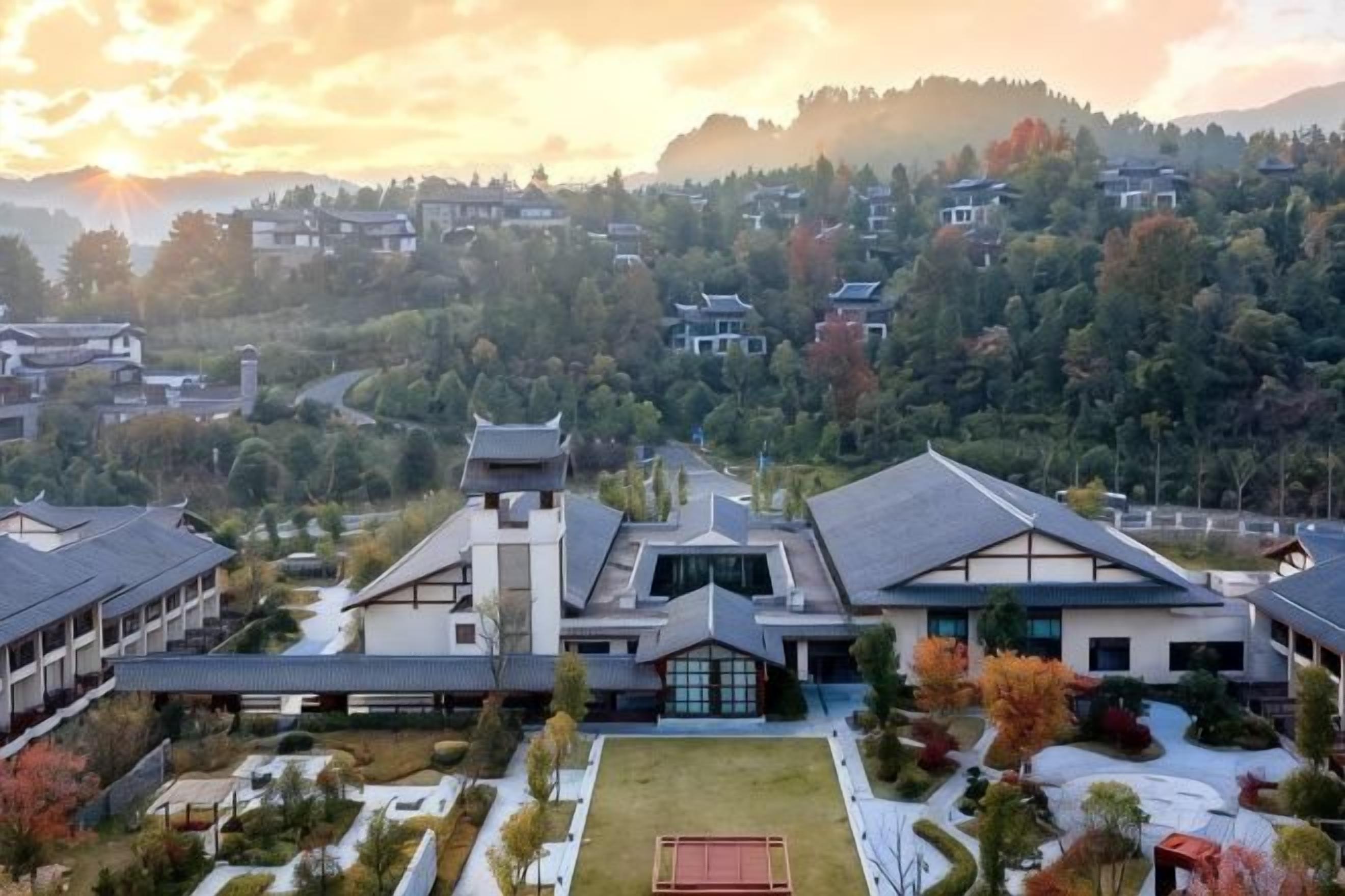 A traditional building with a central tower, surrounded by a courtyard with a tennis court, set against a backdrop of a hilly landscape.