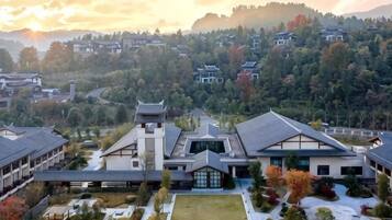 A traditional building with a central tower, surrounded by a courtyard with a tennis court, set against a backdrop of a hilly landscape.
