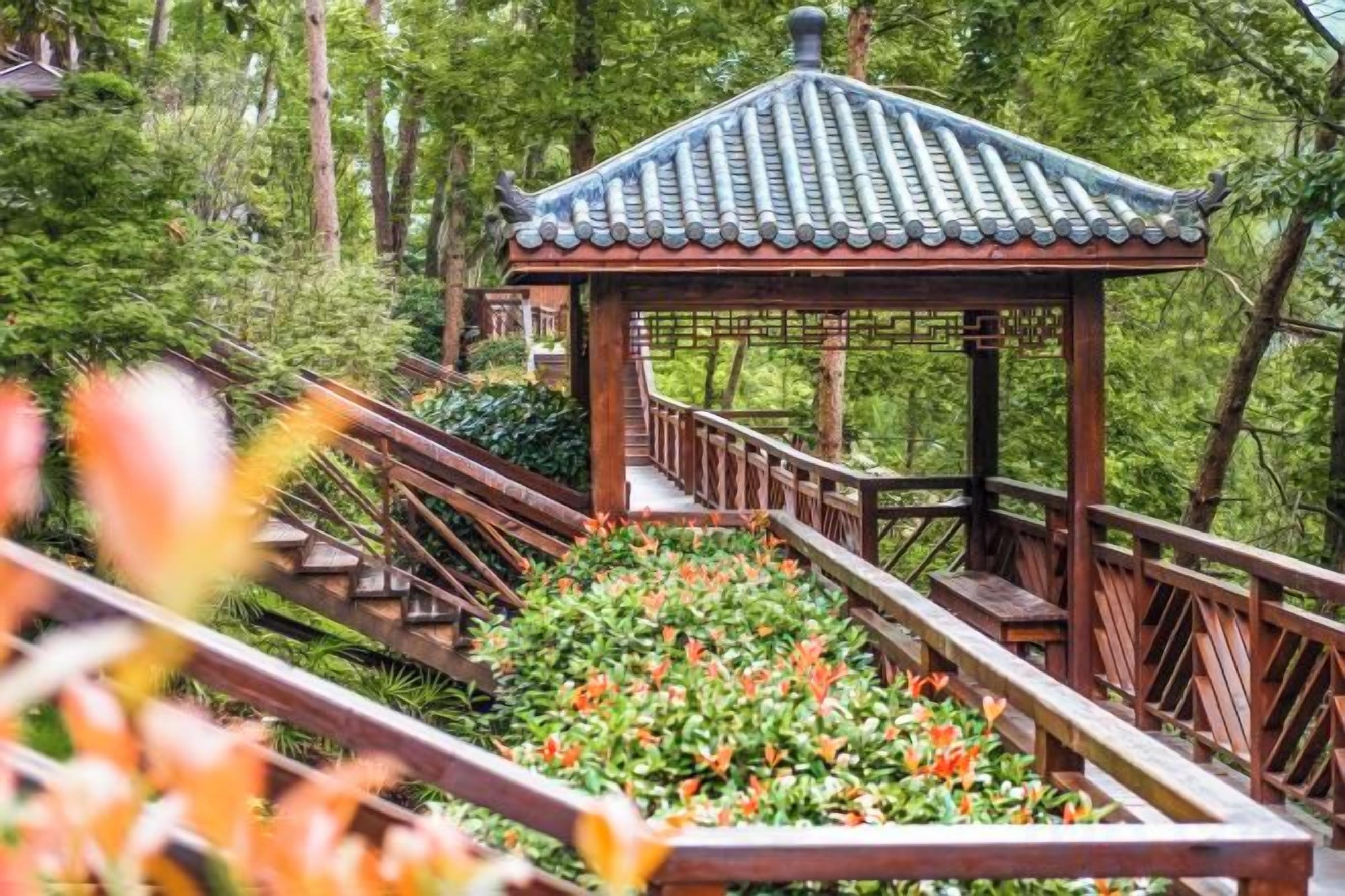 A wooden gazebo with a tiled roof, surrounded by a wooden deck and a garden with flowers.