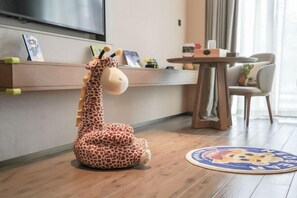Un peluche de jirafa sobre el suelo, una alfombra redonda con personajes de caricatura, una mesa de madera y una silla en una habitación con suelo de madera y un televisor montado en la pared.