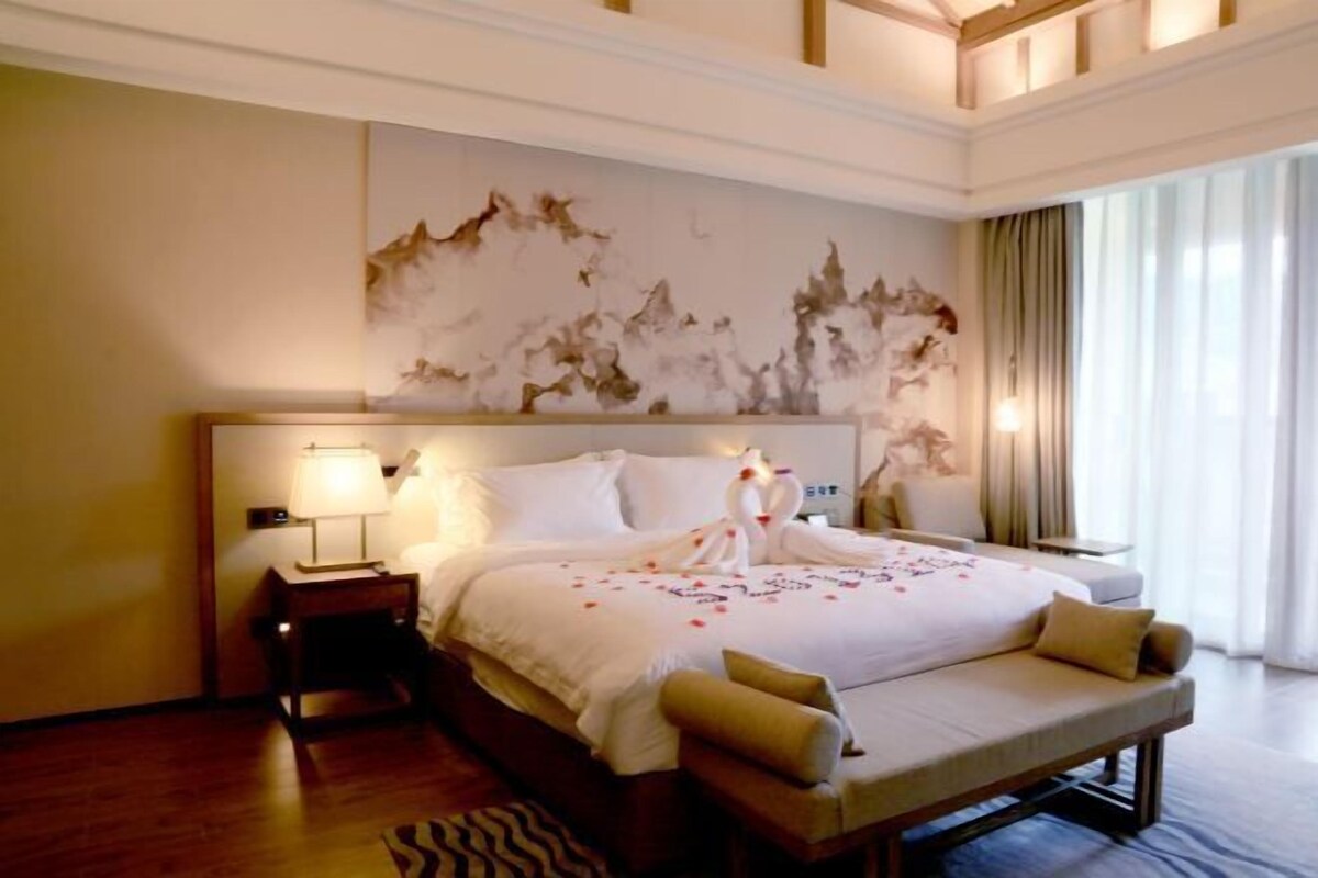 A hotel room with a large bed, a bedside table with a lamp, a decorative wall piece, and a view of the outdoors through a window.