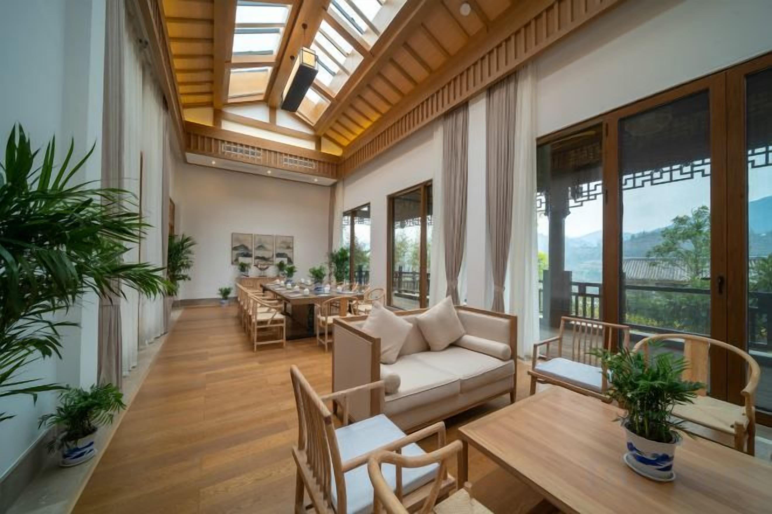 Ein geräumiges Zimmer mit Holzbalkendecke, einer Couch, einem Esstisch und Stühlen. Große Fenster und Kübelpflanzen sind vorhanden.