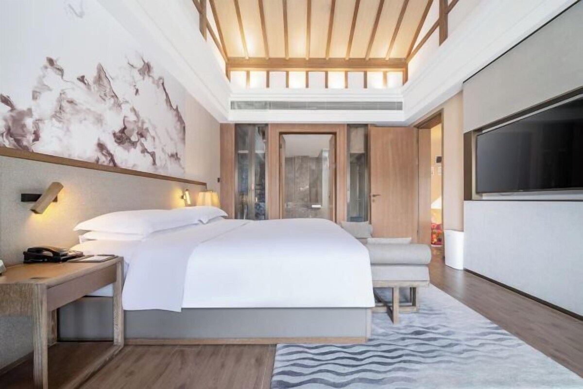 A modern hotel room with a large bed, a desk, a TV, and a bathroom visible through a glass door.