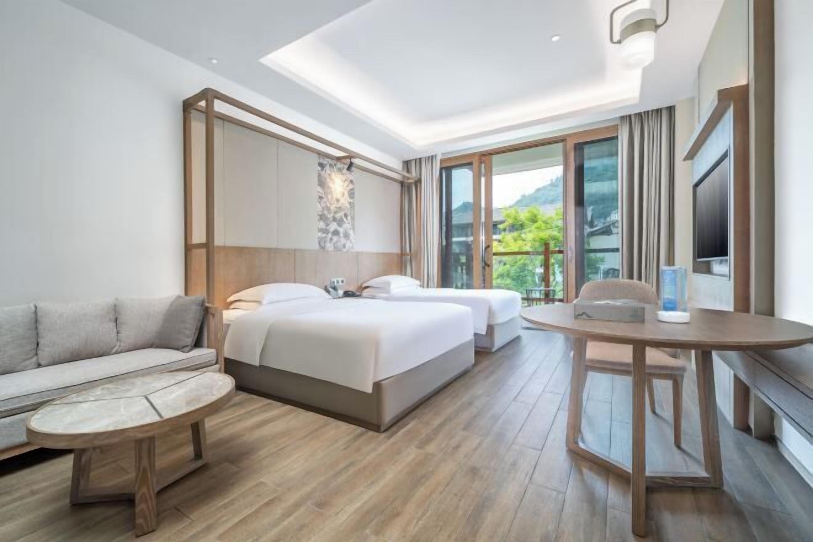 Habitación de hotel moderna con sofá, dos camas, una mesa de comedor y un amplio ventanal con vistas a zonas verdes.