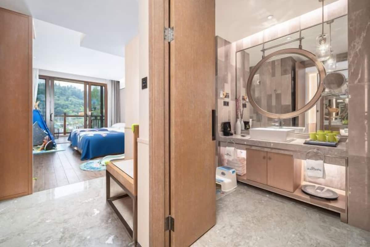 Un baño moderno con un espejo grande, un lavabo y una zona de ducha. Una puerta de madera da paso a un dormitorio con una cama y una tienda de juegos azul.