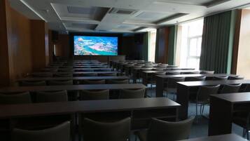 A conference room with rows of tables and chairs, a large screen at the front, and windows with curtains.