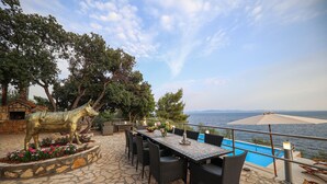 Un patio al aire libre con suelo de piedra, una mesa de comedor con sillas, una estatua de un caballo y una piscina con vistas al mar.