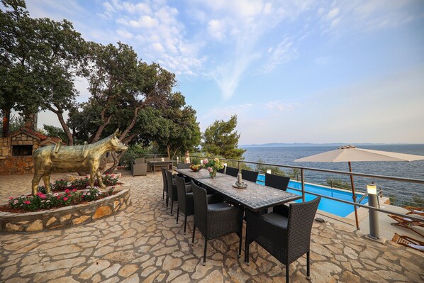 Une terrasse extérieure avec un sol en pierre, une table à manger avec des chaises, une statue de cheval et une piscine offrant une vue sur la mer.