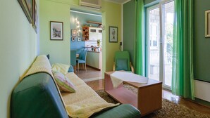 A living room with a green sofa, a small table, and a green chair. There is a kitchen area in the background with a dining table and chairs.