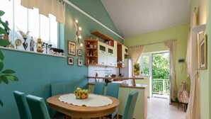 A dining area with a round wooden table and teal chairs, a kitchen with white cabinets, and a view of greenery outside.