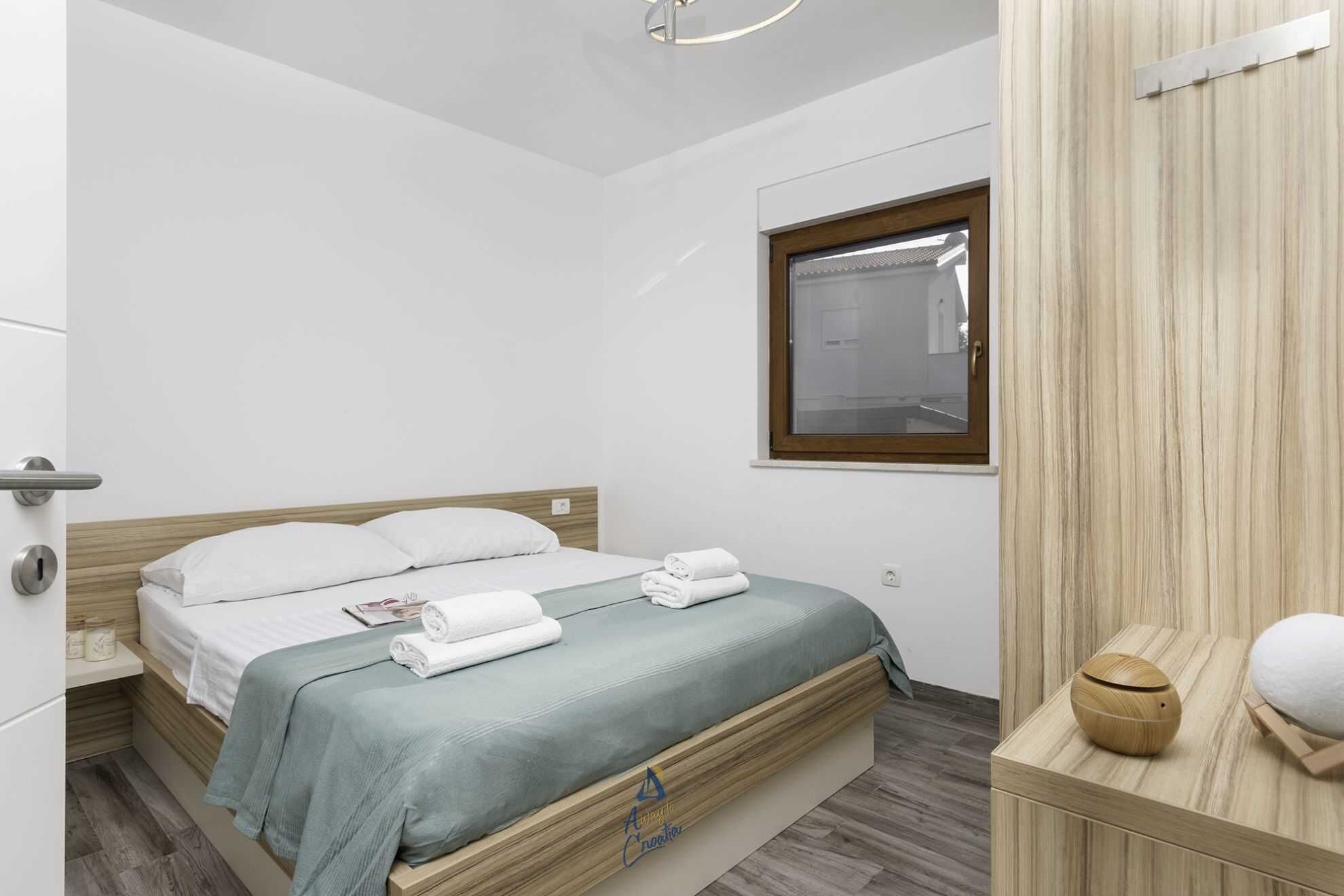 A modern bedroom with a wooden bed, a small window, and a wooden cabinet with a towel and a round object on top.