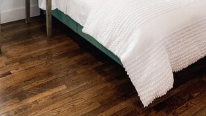 Un dormitorio ordenado con cabecera tapizada en verde, cubrecama blanco y una sola almohada marrón. Un cuadro enmarco cuelga en la pared sobre la cama, y una mesita de noche con una lámpara está situada a la izquierda.