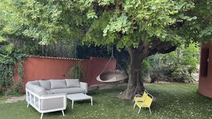 Eine Terrasse mit einem grauen Sofa, einem weißen Tisch und einem gelben Stuhl unter einem Baum.