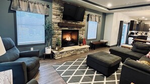A living room with a stone fireplace, a television mounted above it, and a kitchen area visible through an open door. - Private 5 bedroom house in the heart of Laurel highlands! (Normalville)