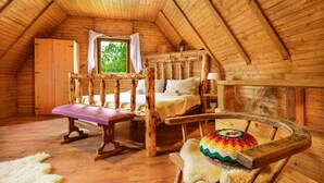 A cozy attic bedroom with a wooden sloped ceiling, a large window with curtains, a wooden bed, a bench, and a colorful woven chair.