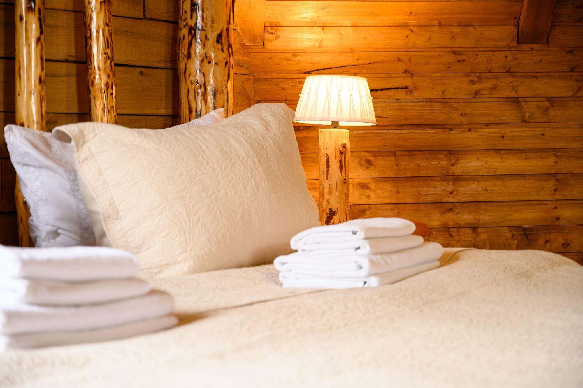 A neatly made bed with white linens and pillows, a stack of folded towels, and a wooden wall with a wooden post.