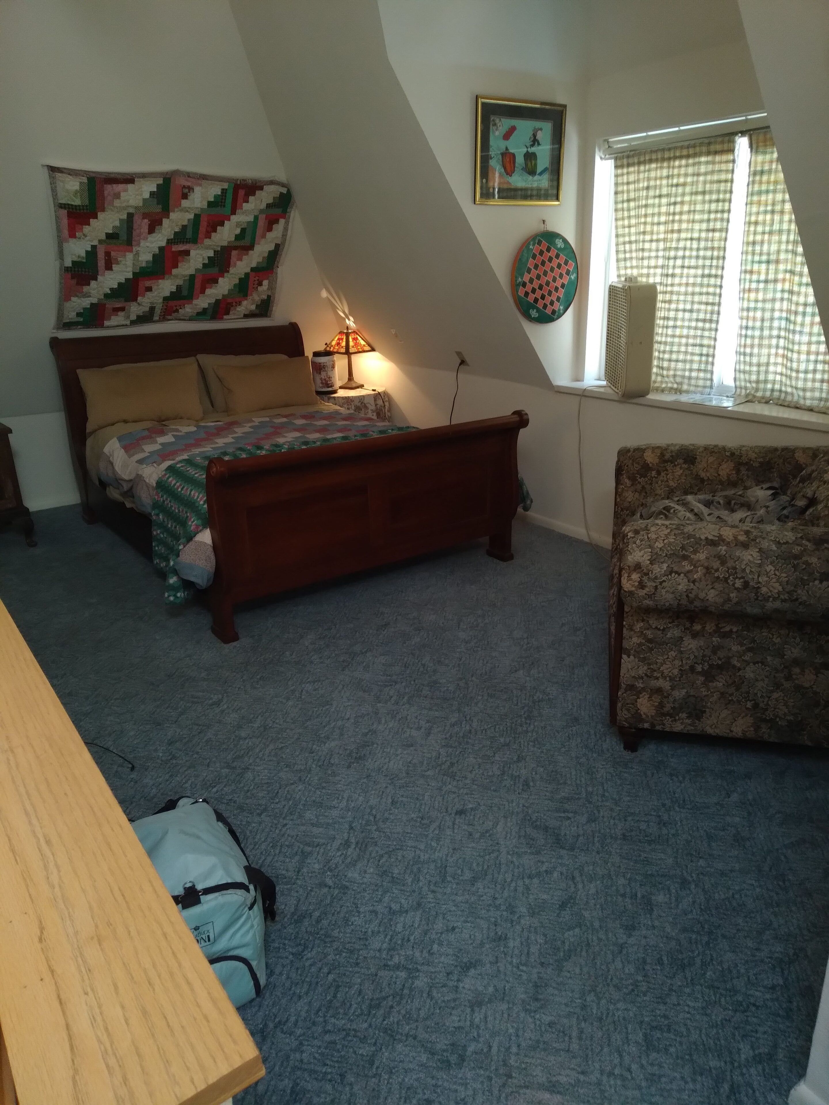 Een slaapkamer met een bed, een houten nachtkastje, een lamp, een sprei, een bloemenbank en een raam met gordijnen.