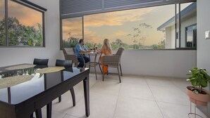 A modern dining area with a glass table and chairs, large windows with a view of trees and a sunset, and a potted plant on the balcony. - La Fortuna Deluxe Vacation Home - 5 Min from Town - Outdoor Pool (Alajuela)