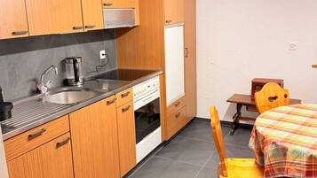 Une cuisine compacte avec des meubles en bois, un évier en acier inoxydable, une machine à café et un petit réfrigérateur. On y trouve une chaise en bois et une table recouverte d’une nappe à carreaux.