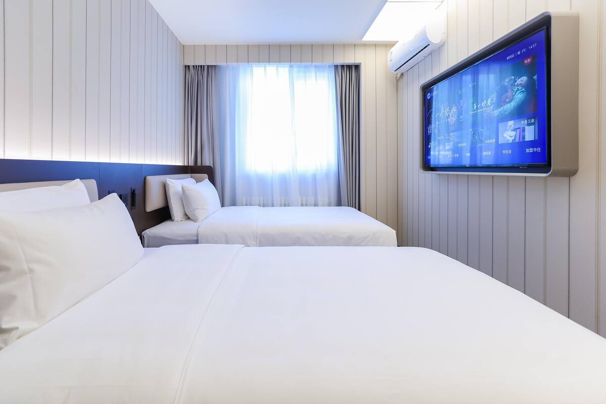 Une chambre d’hôtel avec deux lits, un téléviseur fixé au mur et une fenêtre avec des rideaux.