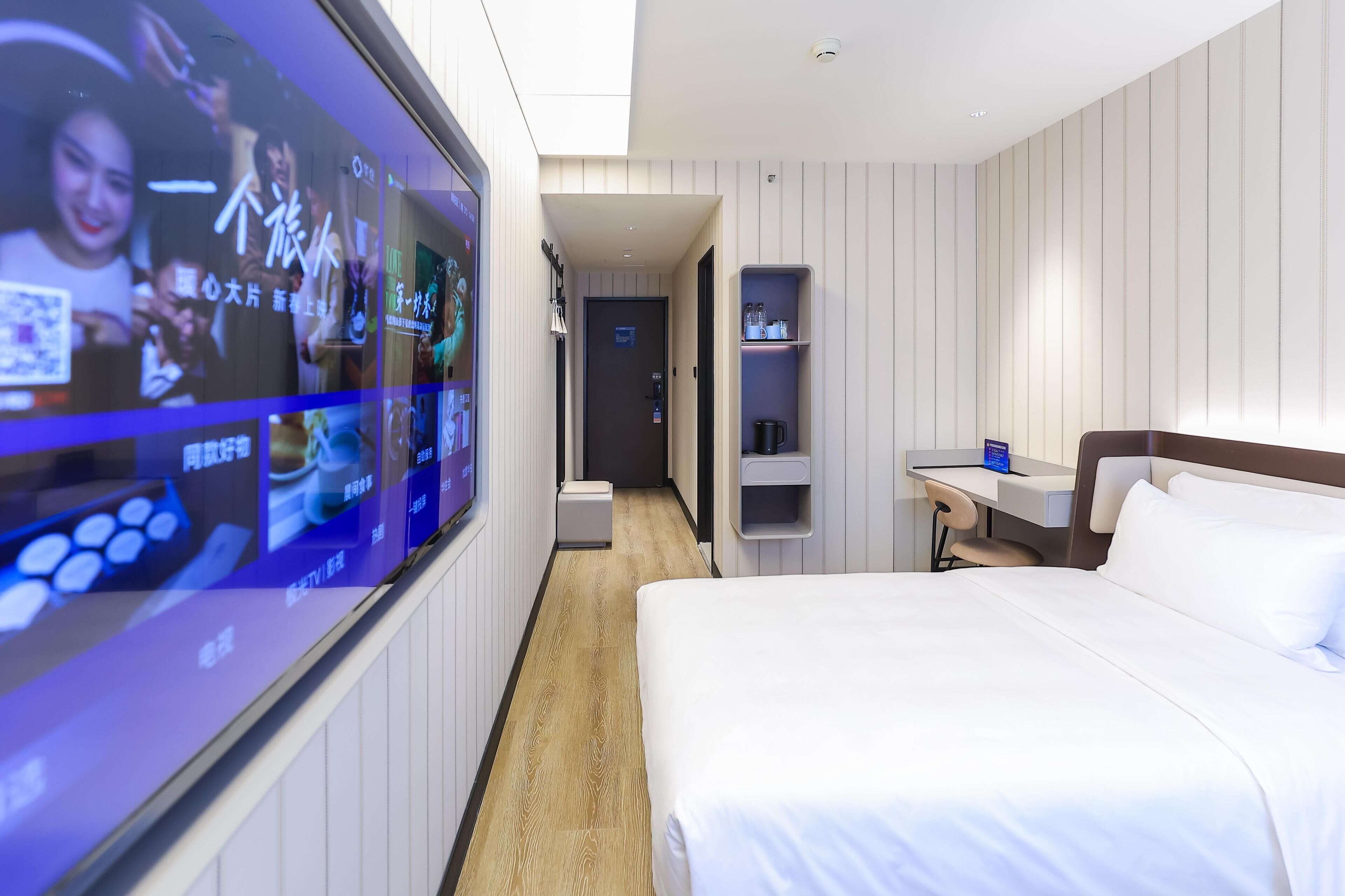 Hotellihuone, jossa on suuri digitaalinen näyttö, sänky, työpöytä ja hyllykkö.