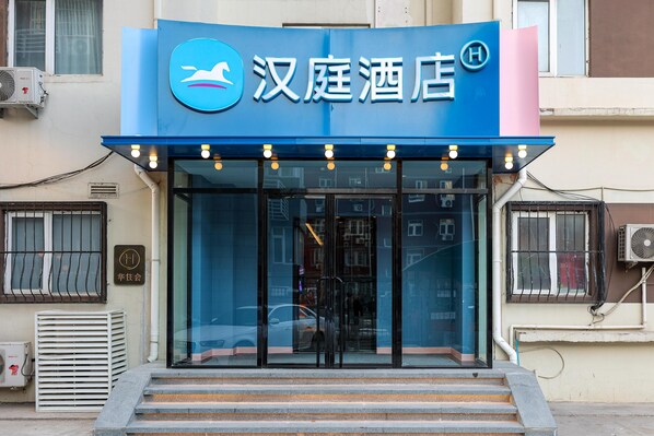 A hotel entrance with a blue awning, glass doors, and a visible air conditioning unit.
