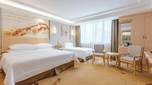 Quarto de hotel com duas camas, uma escrivaninha, uma cadeira e uma janela grande com cortinas.