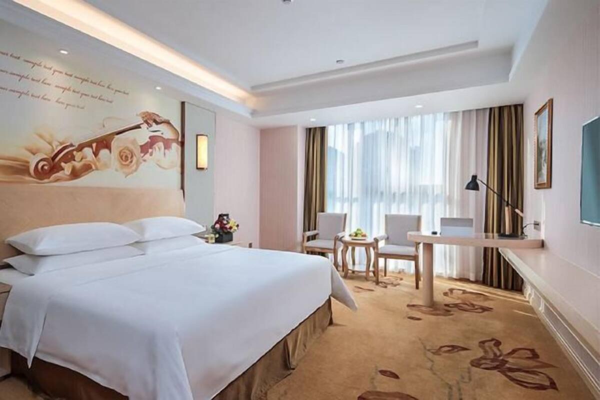 Une chambre d’hôtel avec un grand lit, un bureau, une chaise, une lampe et une fenêtre avec des rideaux.