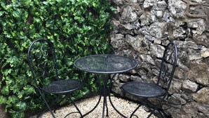 Un'area patio con un tavolo rotondo in metallo e quattro sedie, circondata da un muro in pietra e vegetazione verdeggiante.