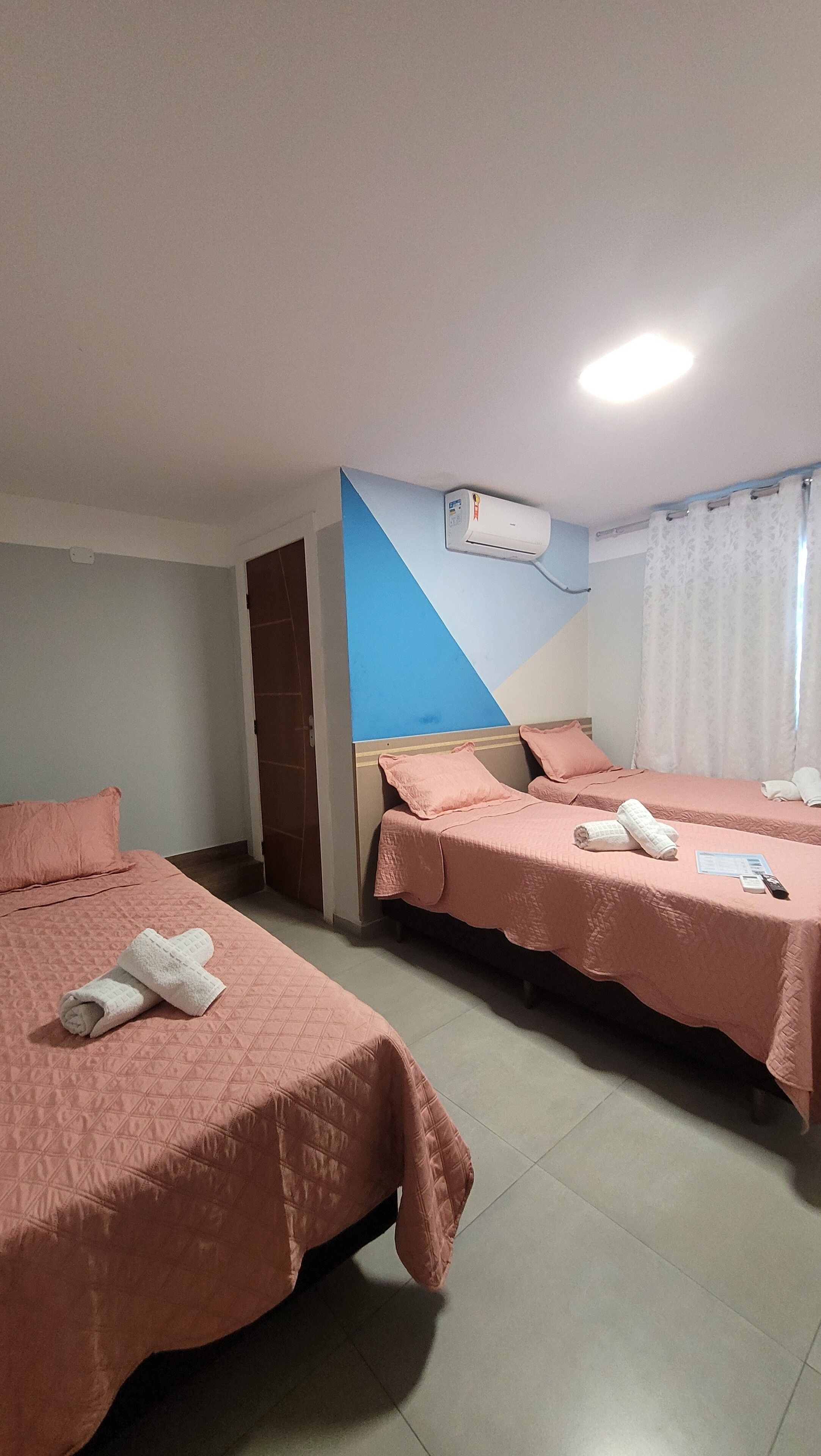 A hotel room with two beds, each with pink bedding and towels. There is a ceiling fan and an air conditioning unit mounted on the wall.