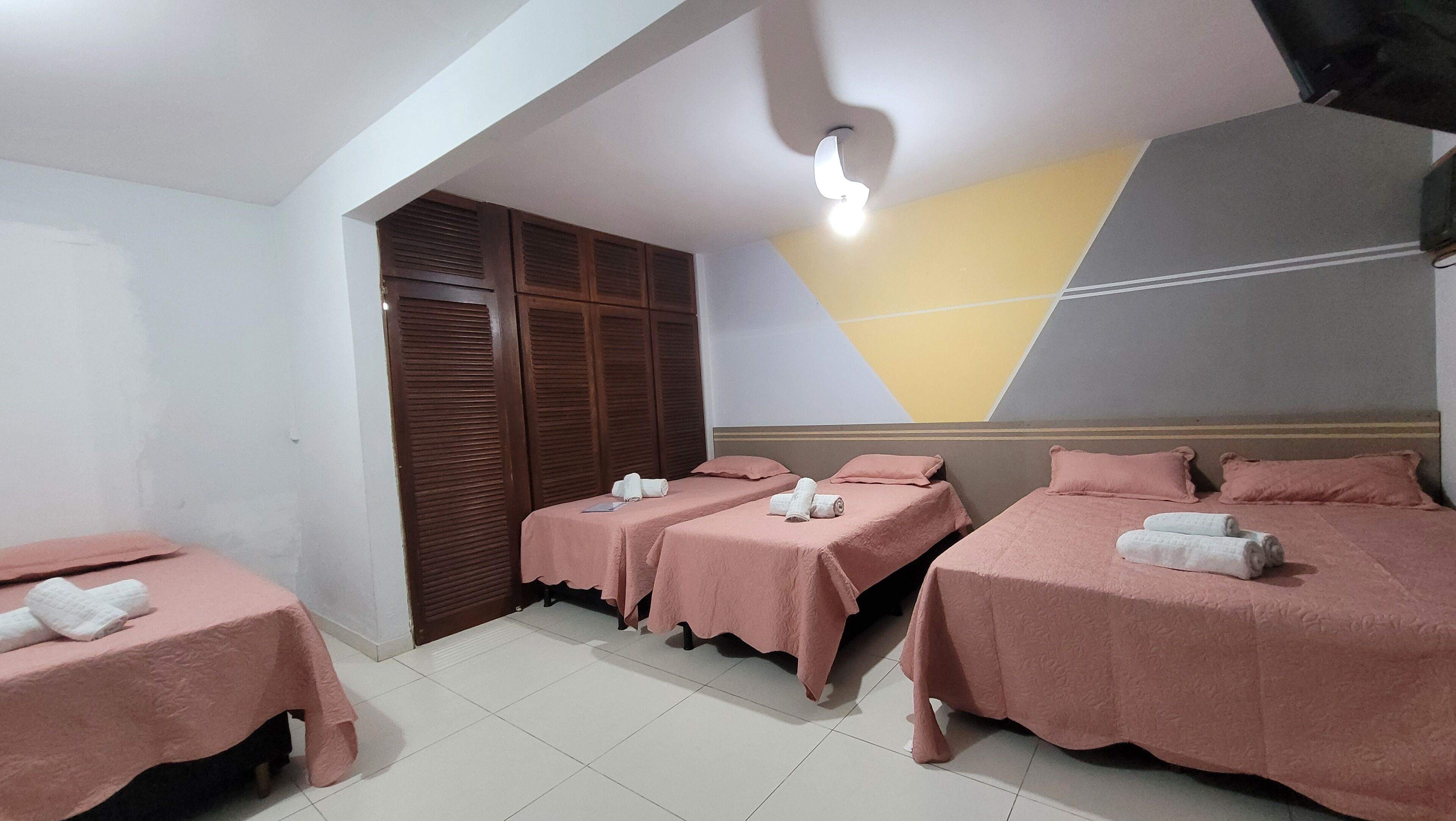 Une chambre d’hôtel avec trois lits, chacun agrémenté de draps et de serviettes roses. On y trouve également une armoire en bois et un luminaire fixé au plafond.