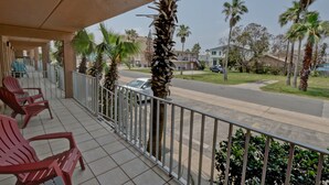 A balcony with red chairs, a view of a street with palm trees, and residential buildings in the background. - Great Condo in the Middle of it ALL! (South Padre Island)