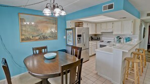 A dining area with a wooden table and chairs, a bowl on the table, and a kitchen in the background with white cabinets and appliances. - Great Condo in the Middle of it ALL! (South Padre Island)