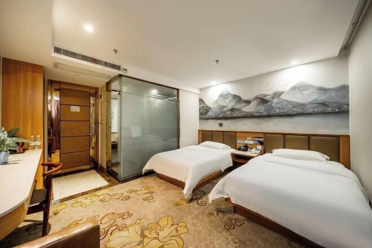 Habitación de hotel con dos camas, un escritorio, una silla, una mesita, un cuadro de montañas y un armario.