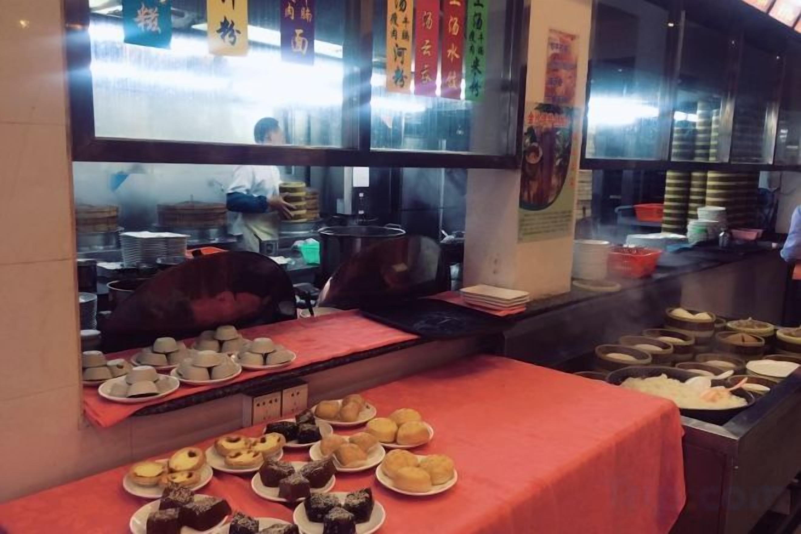 A kitchen area with a table displaying various food items, a window with a view of the street, and kitchen equipment.