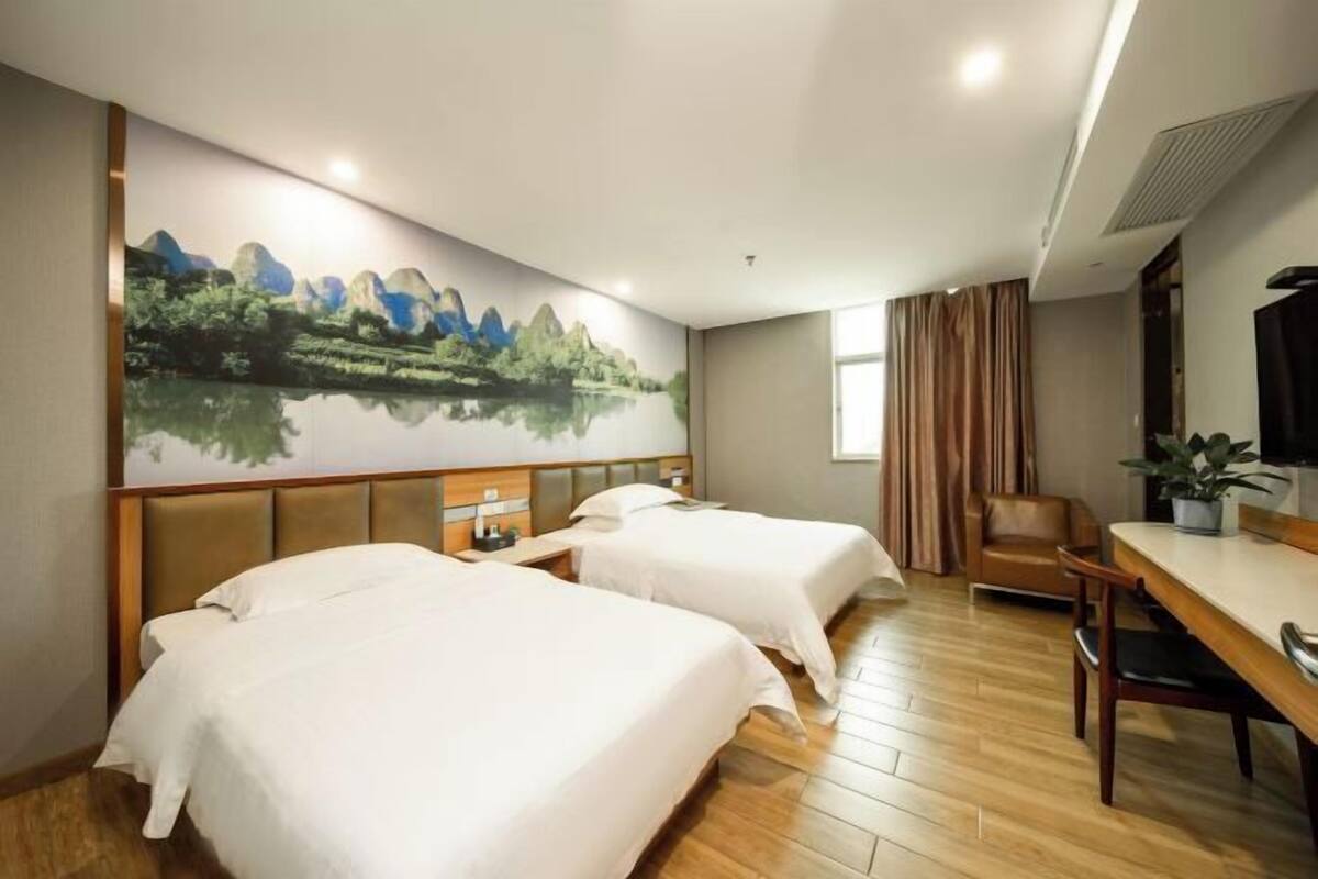 Habitación de hotel con dos camas, una mesa de comedor, una silla, un televisor y un gran mural de un paisaje montañoso.