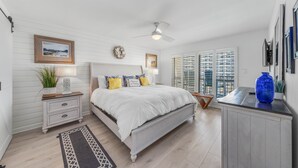 A bedroom with a bed, nightstands, a desk, a large window with blinds, and a ceiling fan.