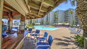 A poolside area with lounge chairs, a bar, and a view of a hotel building.