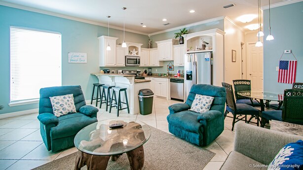 A spacious living room with teal armchairs, a glass coffee table, and a dining area with a table and chairs. A kitchen with white cabinets and a ceiling fan is visible in the background.
