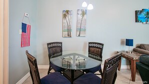 A dining area with a glass table and wicker chairs, a small side table with a lamp, and wall art including an American flag and beach-themed paintings.
