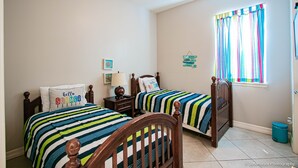 A bedroom with two beds, each with striped bedding and pillows, a nightstand with a lamp, a window with colorful curtains, and a small trash bin.