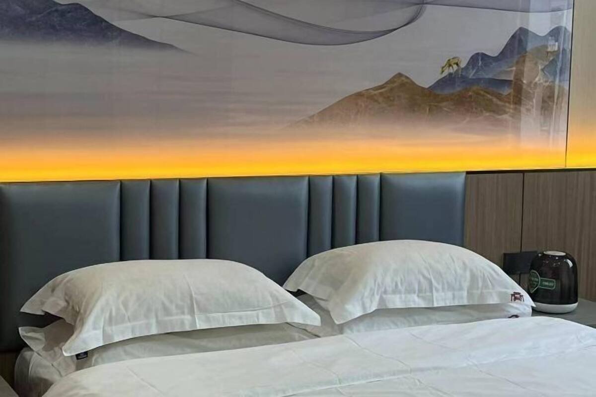 Habitación de hotel con una cama grande, dos almohadas, una mesita de noche con cafetera y un mural en la pared que representa un paisaje montañoso.