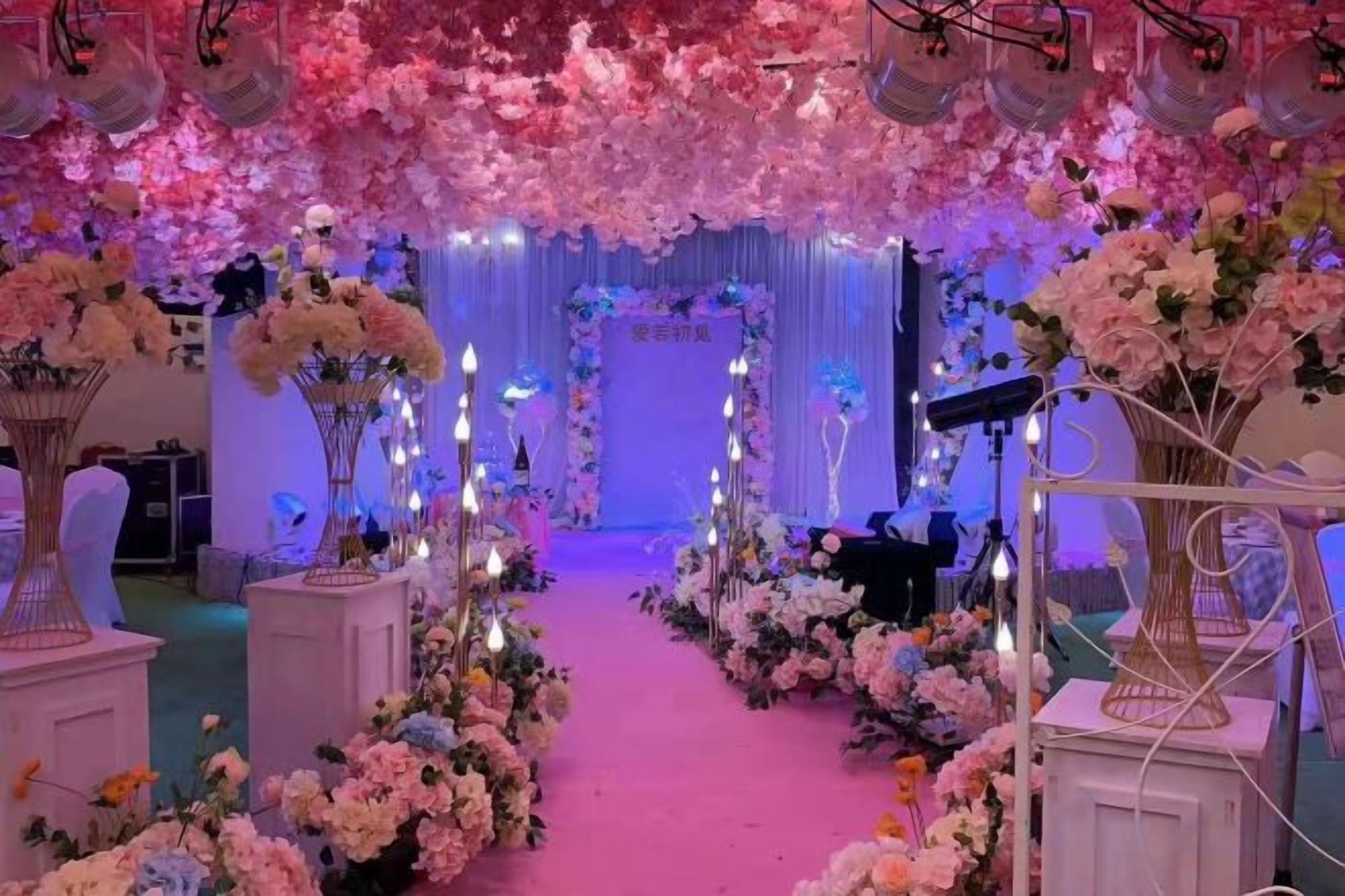 Un pasillo decorado con arreglos florales y velas, que conduce a un escenario con un telón de fondo.