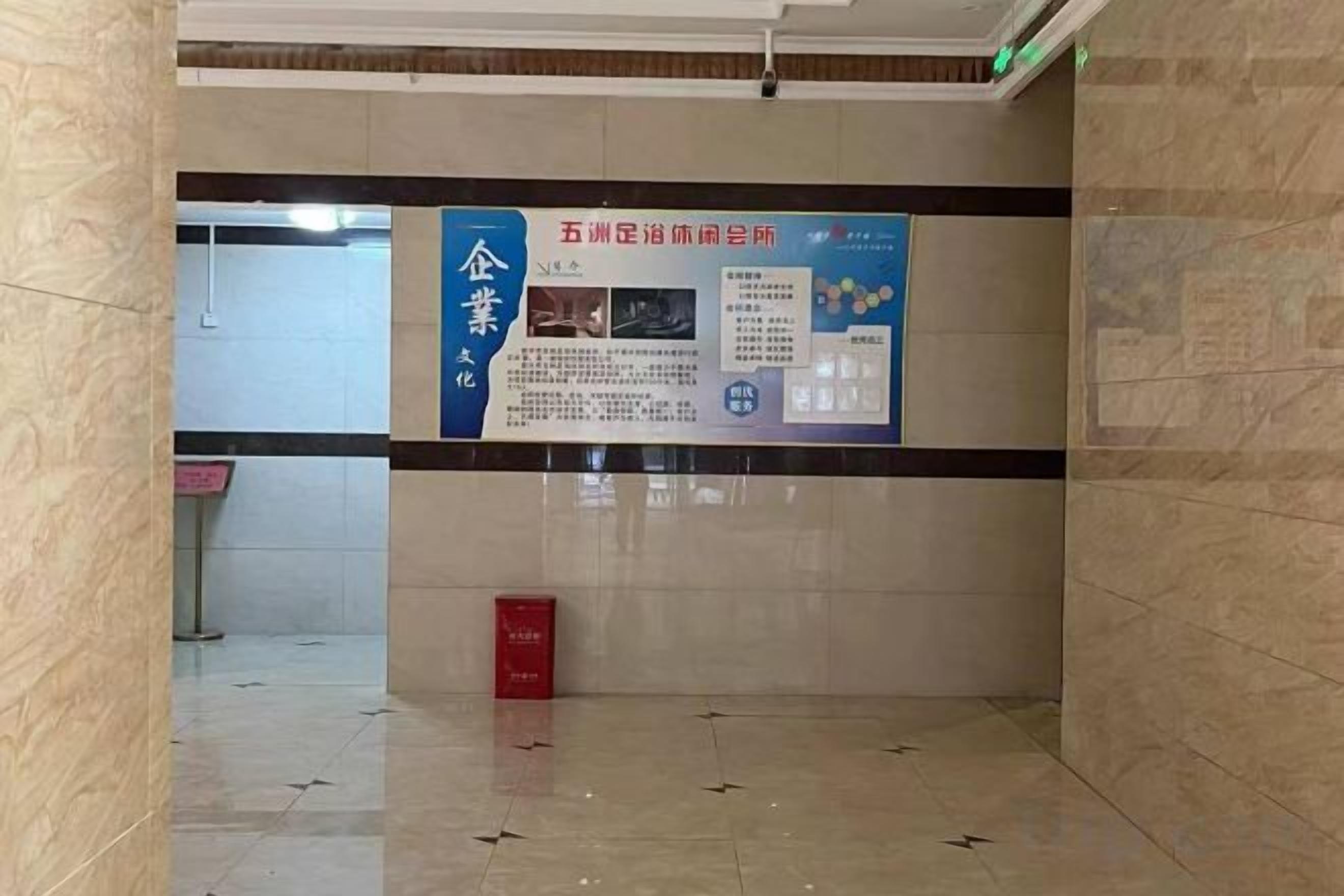 A corridor with a tiled floor, a red trash bin, and a wall with a large informational poster.