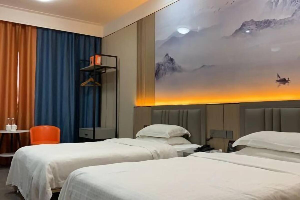 Una habitación de hotel con dos camas, una mesita de noche, una silla, una pequeña mesa y una pared decorada con una pintura de un paisaje de montaña.